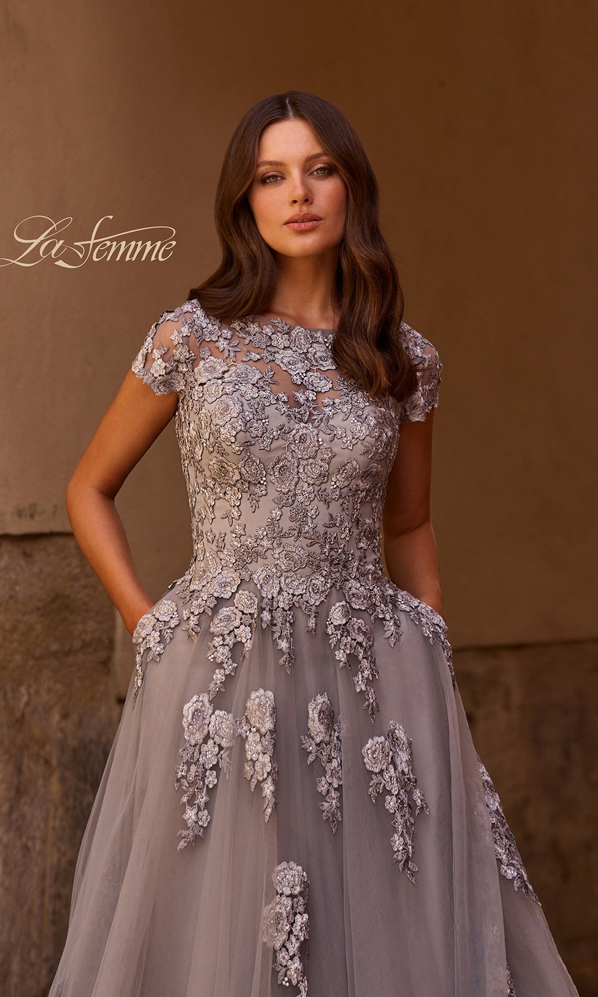 A woman stands against a neutral background, looking at the camera with her hands in the pockets of a gray La Femme 26907 long formal dress. The gown features floral embroidery and embellished lace appliques in an elegant A-line silhouette. “La Femme” is written in script on the left.